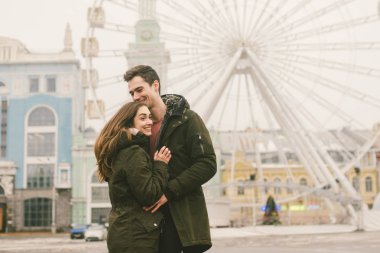 Tema aşk ve tatil Sevgililer günü. Beyaz heteroseksüel aşıklar çifti birlikte kasvetli hava kucaklama çerçevede dönme dolap kasaba Meydanı'nda kış. Adam yavaşça kız hugs.