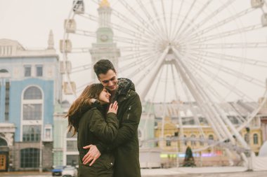Tema aşk ve tatil Sevgililer günü. Beyaz heteroseksüel aşıklar çifti birlikte kasvetli hava kucaklama çerçevede dönme dolap kasaba Meydanı'nda kış. Adam yavaşça kız hugs.