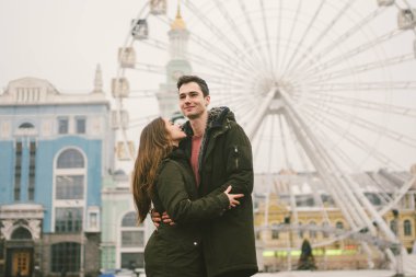 Tema aşk ve tatil Sevgililer günü. Beyaz heteroseksüel aşıklar çifti birlikte kasvetli hava kucaklama çerçevede dönme dolap kasaba Meydanı'nda kış. Adam yavaşça kız hugs.