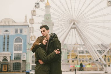 Tema aşk ve tatil Sevgililer günü. Beyaz heteroseksüel aşıklar çifti birlikte kasvetli hava kucaklama çerçevede dönme dolap kasaba Meydanı'nda kış. Adam yavaşça kız hugs.