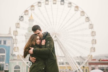 Tema aşk ve tatil Sevgililer günü. Beyaz heteroseksüel aşıklar çifti birlikte kasvetli hava kucaklama çerçevede dönme dolap kasaba Meydanı'nda kış. Adam yavaşça kız hugs.