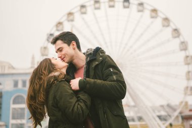 Tema aşk ve tatil Sevgililer günü. Beyaz heteroseksüel aşıklar çifti birlikte kasvetli hava kucaklama çerçevede dönme dolap kasaba Meydanı'nda kış. Adam yavaşça kız hugs.