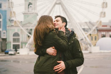 Tema aşk ve tatil Sevgililer günü. Beyaz heteroseksüel aşıklar çifti birlikte kasvetli hava kucaklama çerçevede dönme dolap kasaba Meydanı'nda kış. Adam yavaşça kız hugs.
