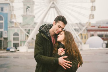 Tema aşk ve tatil Sevgililer günü. Beyaz heteroseksüel aşıklar çifti birlikte kasvetli hava kucaklama çerçevede dönme dolap kasaba Meydanı'nda kış. Adam yavaşça kız hugs.