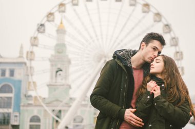 Tema aşk ve tatil Sevgililer günü. Beyaz heteroseksüel aşıklar çifti birlikte kasvetli hava kucaklama çerçevede dönme dolap kasaba Meydanı'nda kış. Adam yavaşça kız hugs.