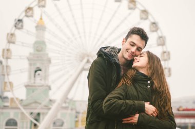 Tema aşk ve tatil Sevgililer günü. Beyaz heteroseksüel aşıklar çifti birlikte kasvetli hava kucaklama çerçevede dönme dolap kasaba Meydanı'nda kış. Adam yavaşça kız hugs.