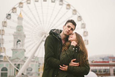 Tema aşk ve tatil Sevgililer günü. Beyaz heteroseksüel aşıklar çifti birlikte kasvetli hava kucaklama çerçevede dönme dolap kasaba Meydanı'nda kış. Adam yavaşça kız hugs.