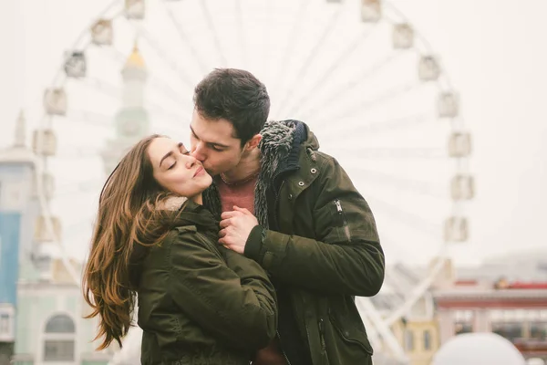 Theme love and holiday Valentines Day. pair of Caucasian heterosexual lovers in winter together gloomy weather embrace against background of Ferris wheel in town square. The guy gently hugs the girl.