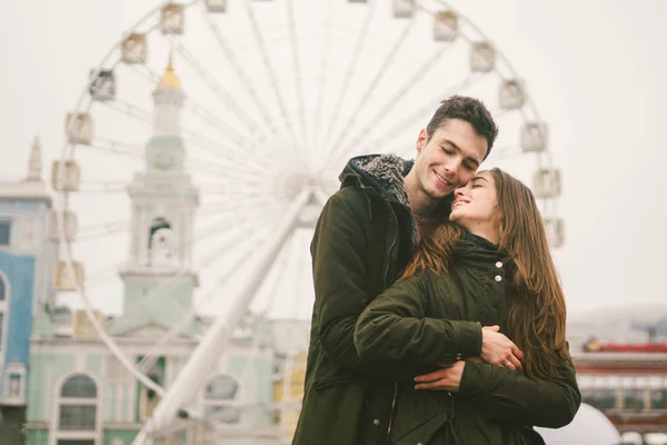 Theme love and holiday Valentines Day. pair of Caucasian heterosexual lovers in winter together gloomy weather embrace against background of Ferris wheel in town square. The guy gently hugs the girl.