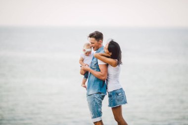 Doğada aile tatili. Üç, anne, baba, kızını bir yıl denize bakan cliff sırtında ile ayakta. kollarında, kadın kucaklama koca Hild tutan adam. Şık giysiler giymiş şık insanlar.