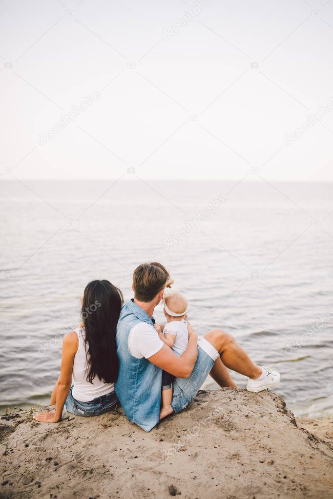 Theme family vacation with small child on the nature and sea. Mom, Dad and daughter of one year are sitting in embrace, girls in the hands of man, with his back at height of cliff overlooking the sea.