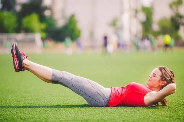 Tema deporte y salud. Joven hermosa mujer caucásica haciendo calentamiento, el calentamiento de los músculos, ejercicios musculares abdominales, la pérdida de vientre, la hierba verde abdominal en el estadio en el verano en césped artificial — Foto de Stock