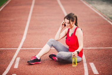 Spor ve teknoloji. At kuyruğu oturma Stadyumu'nda, kırmızı koşu bandı parça çalışma sırasında egzersiz yaptıktan sonra dinlenme ile bir güzel genç beyaz bir kadın. Cep telefonu kulaklık ve spor saatler kullanımları