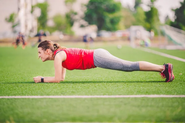 Theme sport and health. Young Caucasian woman doing warm-up, warming up muscles, training abdominal muscles. Losing belly. Abdominal plank exercise on green grass in stadium summer artificial turf