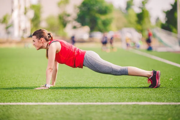 Tema esporte e saúde. Mulher branca jovem fazendo aquecimento, aquecendo músculos, treinando músculos abdominais. Perder a barriga. Exercício de prancha abdominal na grama verde no estádio de verão relva artificial — Fotografia de Stock