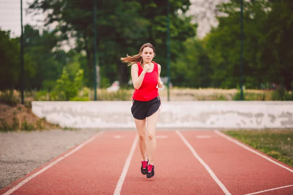 Όμορφη νεαρός αθλητής καυκάσιος γυναίκα με μεγάλα στήθη σε κόκκινο T-shirt και κοντά σορτς τρέξιμο, τρέξιμο στο γήπεδο με κόκκινο καουτσούκ επίστρωση — Φωτογραφία Αρχείου