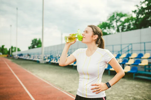 Theme is sport and health. Beautiful young caucasian woman with big breasts athlete runner stands resting on running stadium, running track with bottle in hands drinking water in short shorts