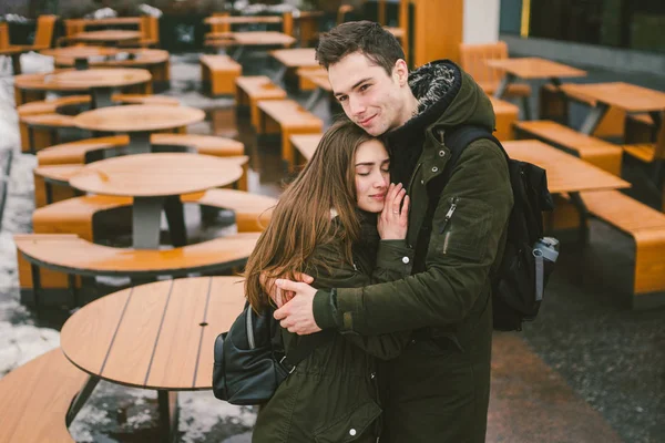 Una giovane coppia innamorata e una ragazza e uno studente si alzano abbracciati vicino ai tavoli di una terrazza di strada bar chiuso vuoto senza persone per l'inverno. Fuori stagione ristorazione ristorante brutto tempo — Foto Stock