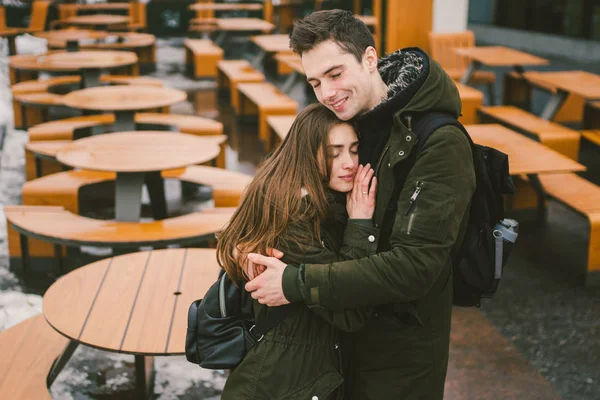 Um jovem casal apaixonado e uma menina e um estande estudantil abraçando perto das mesas de um café terraço de rua fechado vazio sem pessoas para o inverno. Off-temporada restaurante de catering mau tempo — Fotografia de Stock