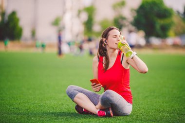 Tema spor sağlık. güzel genç kız oturan yeşil çimenlerin üzerinde dinlenme. çim Stadı technodogies kullanarak. Handphone kulak kulaklık müzik, yaz spor giyim akıllı seyretmek kalp oran sınıf başkanı dinler