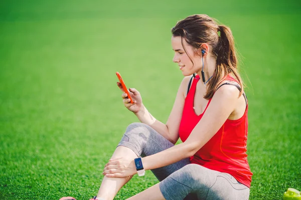 主题运动健康 美丽的年轻女孩坐在绿草上休息 使用技术的草坪体育场 在手机耳机听音乐 夏季运动服装智能手表心率监测仪 — 图库照片