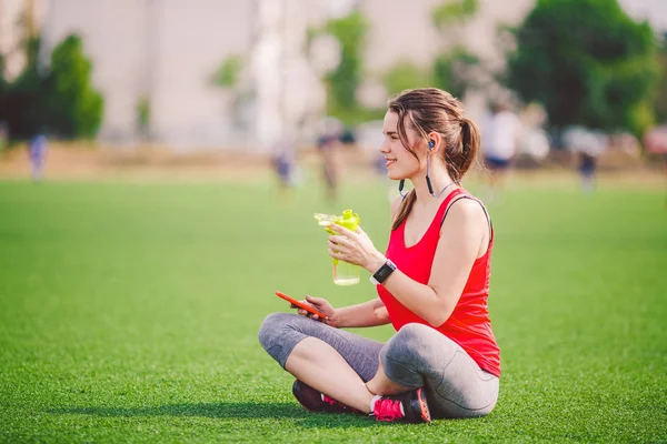主题运动健康 美丽的年轻女孩坐在绿草上休息 使用技术的草坪体育场 在手机耳机听音乐 夏季运动服装智能手表心率监测仪 — 图库照片