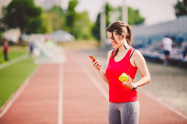 Tema salud deportiva. chica de pie descansando en la pista. carretera del estadio utiliza tecnodogies. auriculares para el oído del teléfono de la mano escucha música. Reloj inteligente de mano de ropa deportiva de verano monitor de frecuencia cardíaca, bebida botella de agua — Foto de Stock