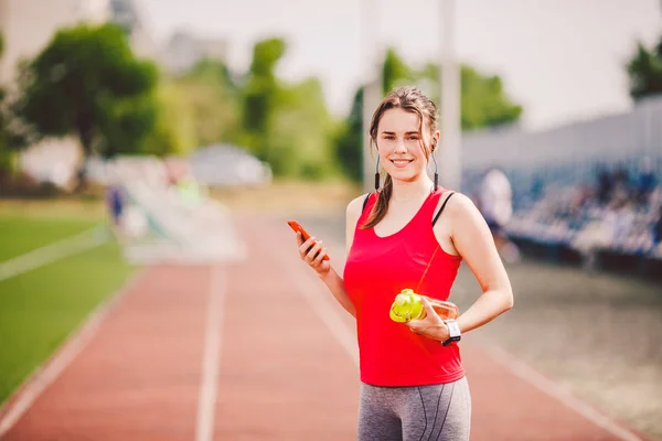 Tema salud deportiva. chica de pie descansando en la pista. carretera del estadio utiliza tecnodogies. auriculares para el oído del teléfono de la mano escucha música. Reloj inteligente de mano de ropa deportiva de verano monitor de frecuencia cardíaca, bebida botella de agua — Foto de Stock