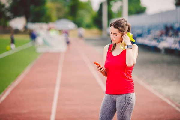 Tema salud deportiva. chica de pie descansando en la pista. carretera del estadio utiliza tecnodogies. auriculares para el oído del teléfono de la mano escucha música. Reloj inteligente de mano de ropa deportiva de verano monitor de frecuencia cardíaca, bebida botella de agua — Foto de Stock