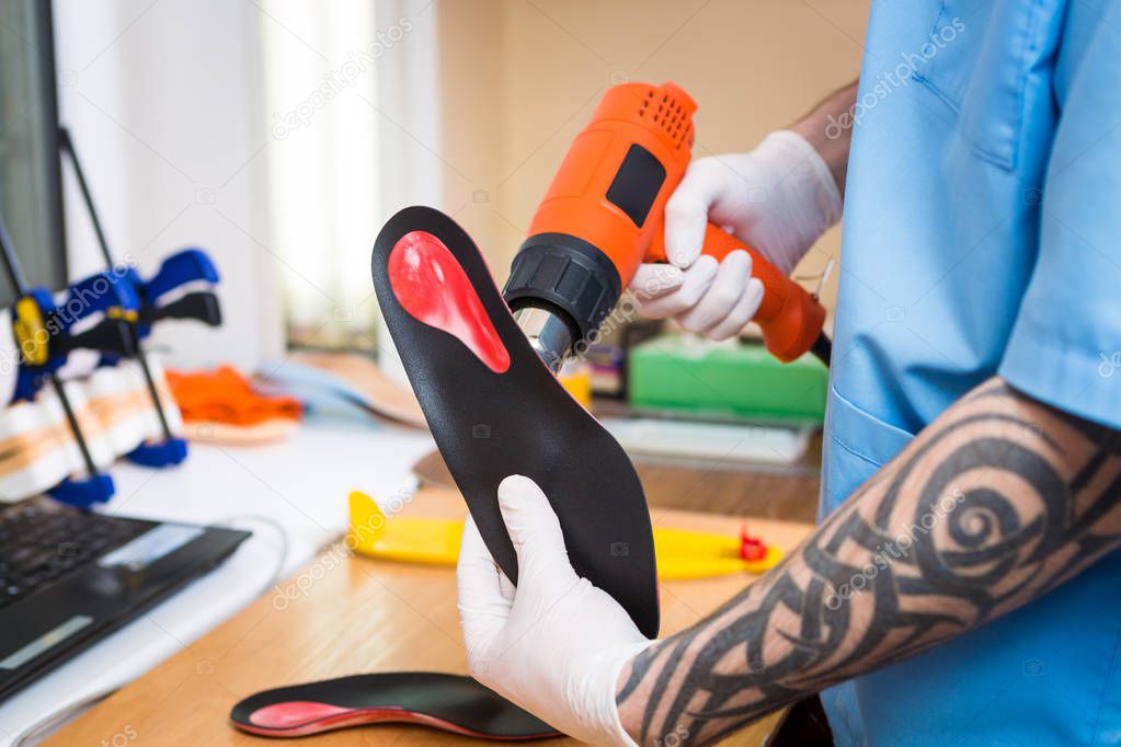 closeup Hands of young man with tattoo in workshop dressed in blue uniform make individual orthopedic insoles. The instrument uses hairdryer to heat and deform. Theme of small business and medicine.