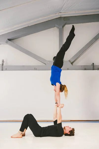 Tema é esportes e área. Um jovem casal de homens e mulheres caucasianos praticando ioga acrobática em um ginásio branco em esteiras. Um homem deita-se de costas e segura uma mulher de braços estendidos. Dose HAND-TO-HAND — Fotografia de Stock