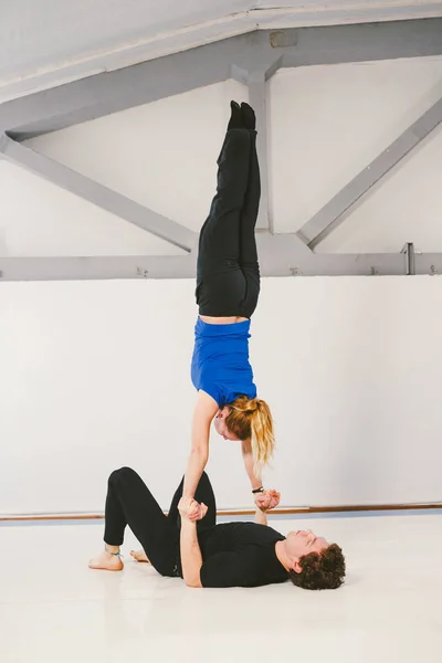 Tema é esportes e área. Um jovem casal de homens e mulheres caucasianos praticando ioga acrobática em um ginásio branco em esteiras. Um homem deita-se de costas e segura uma mulher de braços estendidos. Dose HAND-TO-HAND — Fotografia de Stock