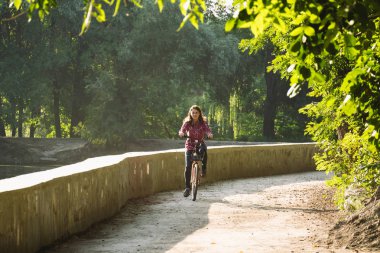 Konu ekolojik bisiklet taşıma. Beyaz genç bir kadın bir toprak yola yakın bir göl güneşli havalarda sonbaharda bir turuncu renkli Bisiklet Kiralama bir parkta sürme.
