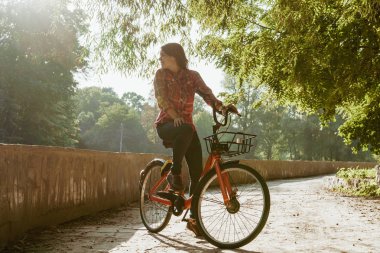 Konu ekolojik bisiklet taşıma. Güneşli havalarda sonbaharda bir dinlenme için poz bir turuncu renkli Bisiklet Kiralama bir Gölü yakınındaki bir parkta bir toprak yol üzerinde genç beyaz kadın.