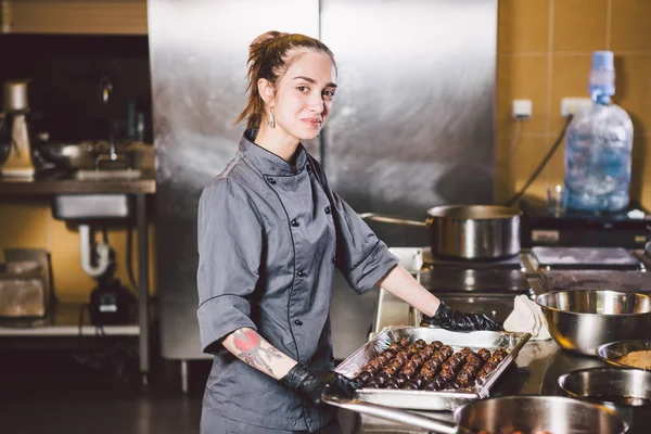 Subject profession and cooking pastry. young Caucasian woman with tattoo of pastry chef in kitchen of restaurant preparing round chocolate candies handmade truffle in black gloves and uniform.