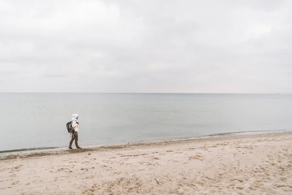 Un hombre caucásico joven turista caucásico con una mochila negra en una playa de arena cerca del mar Báltico en invierno. Viaje temático solo. Pensamientos y sueños con vistas al horizonte —  Fotos de Stock