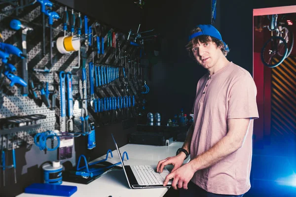 Tema Gerente de profesión empresarial y tecnología. Joven elegante caucásico propietario tienda de bicicletas utiliza una computadora portátil cerca del stand con las herramientas para reparar y dar servicio a la bicicleta —  Fotos de Stock