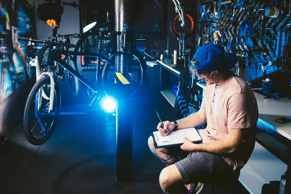 잘생긴 세련 된 젊은이 모자 snapback와 자전거를 판매 하는 문신 중소 기업 소유자, 워크샵 메모를 작성 하는 노트북의 손에 저장소의 백그라운드에 앉아 — 스톡 사진