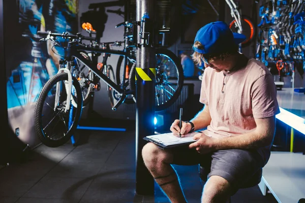 Joven hombre guapo con estilo en un snapback gorra y con un pequeño empresario tatuaje propietario de la venta de una bicicleta, el taller se sienta en el fondo de la tienda en las manos de un cuaderno para escribir notas —  Fotos de Stock