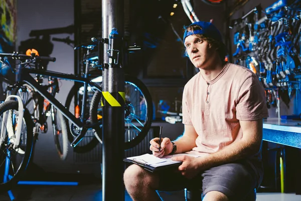 Joven hombre guapo con estilo en un snapback gorra y con un pequeño empresario tatuaje propietario de la venta de una bicicleta, el taller se sienta en el fondo de la tienda en las manos de un cuaderno para escribir notas —  Fotos de Stock