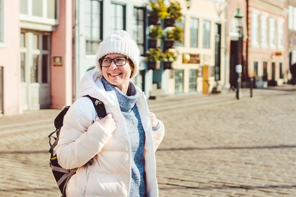 Retrato de uma jovem mulher europeia caucasiana turista de óculos para a vista de um chapéu branco e uma jaqueta com uma mochila preta posando em uma antiga rua da Europa na cidade de Copenhague, na Dinamarca — Fotografia de Stock