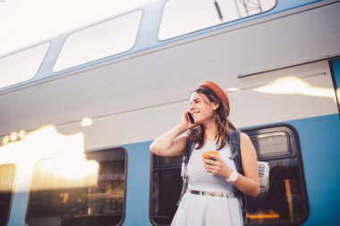 Tema turizm ve seyahat genç öğrenci. güzel genç beyaz kız elbise ve şapka tren istasyonu yakınındaki tren sırt çantası ve fincan kahve ile el ayakta. Smartphone telefon teknolojisini kullanır