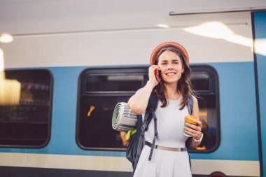 Tema turizm ve seyahat genç öğrenci. güzel genç beyaz kız elbise ve şapka tren istasyonu yakınındaki tren sırt çantası ve fincan kahve ile el ayakta. Smartphone telefon teknolojisini kullanır