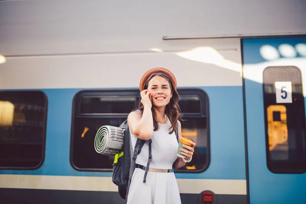 Turismo temático e viagens jovem estudante. bela menina caucasiana em vestido e chapéu de pé na estação de trem perto de trem com mochila e xícara de café na mão. Usa a tecnologia de telefone smartphone — Fotografia de Stock