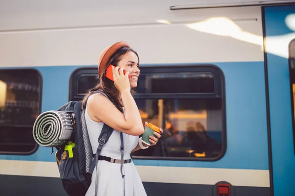 Turismo temático e viagens jovem estudante. bela menina caucasiana em vestido e chapéu de pé na estação de trem perto de trem com mochila e xícara de café na mão. Usa a tecnologia de telefone smartphone — Fotografia de Stock