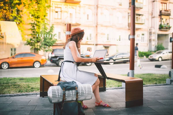 Turismo temático y viajar joven estudiante. hermosa chica caucásica joven en vestido y sombrero se sienta en la cafetería de la calle en la mesa de madera utilizando la tecnología de computadoras portátiles y teléfono móvil. Buscar y reservar hotel y billete — Foto de Stock