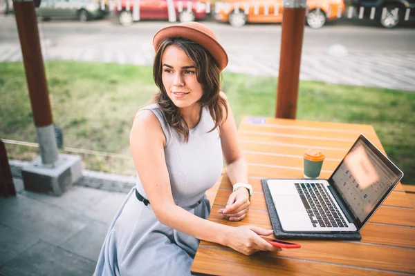 Turismo temático e viagens jovem estudante. bela jovem caucasiana menina em vestido e chapéu senta-se no café rua à mesa de madeira usando a tecnologia laptop e telefone móvel. Pesquisar e reservar hotel e bilhete — Fotografia de Stock