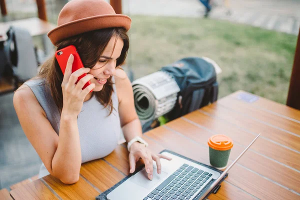 Turismo tematico e viaggi giovane studente. bella giovane ragazza caucasica in abito e cappello si siede in caffè di strada a tavola di legno utilizzando la tecnologia portatile e il telefono cellulare. Cerca e prenota hotel e biglietti — Foto Stock