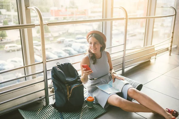 Тема туризму та подорожей молодого студента. Красива молода біла дівчина в одязі і капелюсі сидить на підлозі туристичного килима всередині терміналу аеропорту. Кімната очікування затримала рейс, затримка відправлення — стокове фото