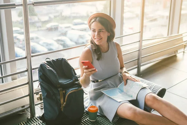 Tema del turismo y los viajes de los jóvenes estudiantes. Hermosa chica caucásica joven en vestido y sombrero se sienta en la alfombra turística piso dentro de la terminal terminal del aeropuerto. Sala de espera vuelo retrasado, salida retrasada —  Fotos de Stock